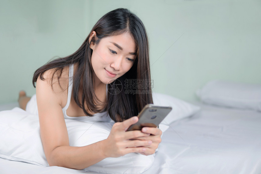 可爱的亚洲女人在卧室和微笑中使用手机图片
