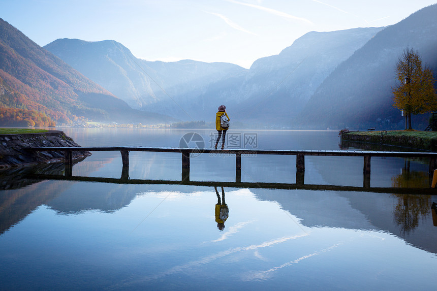 带着帽子和背包的女游客清晨站在山湖上的木桥美丽风景和反光图片
