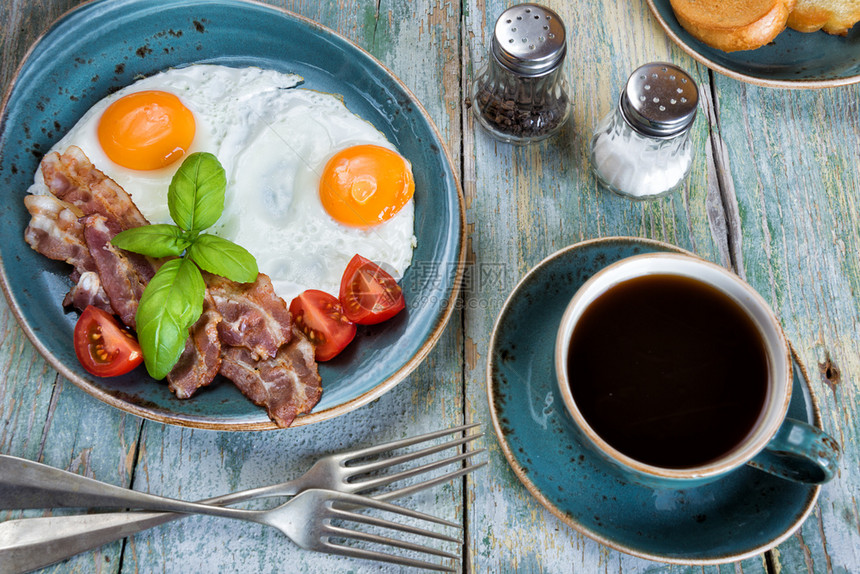 早餐包括炸鸡蛋培根生番茄在旧蓝色瓷板盘上烤面包和在旧木制桌上的黑咖啡图片