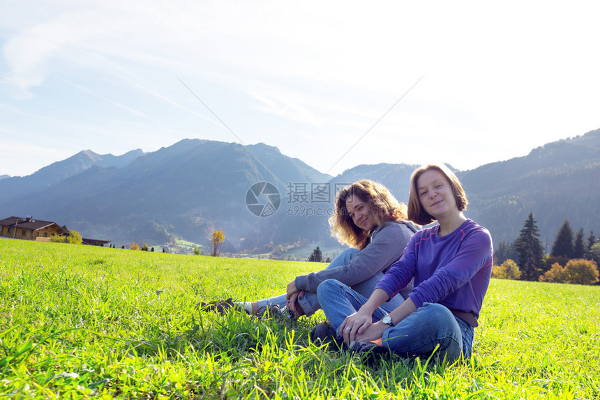 两个微笑的女孩坐在绿色草地上和山的背景图片