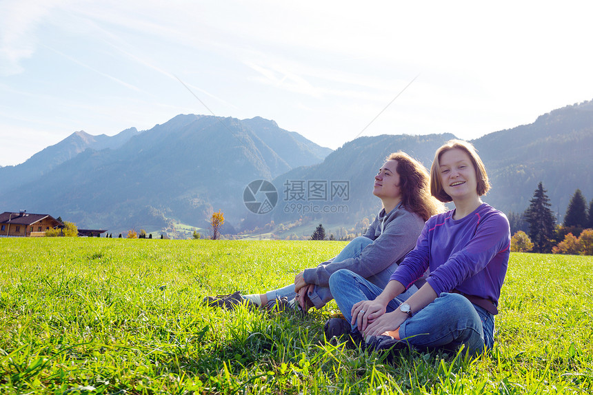 两个微笑的女孩坐在绿色草地上和山的背景图片