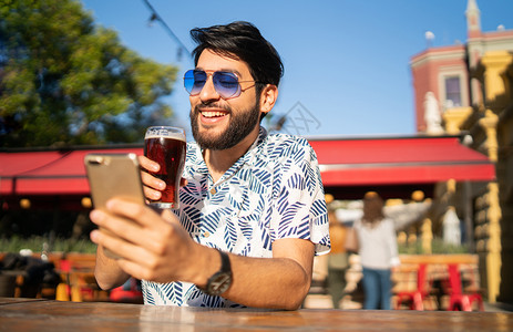 英俊的年轻男子享受夏季天气使用移动电话在户外喝啤酒图片