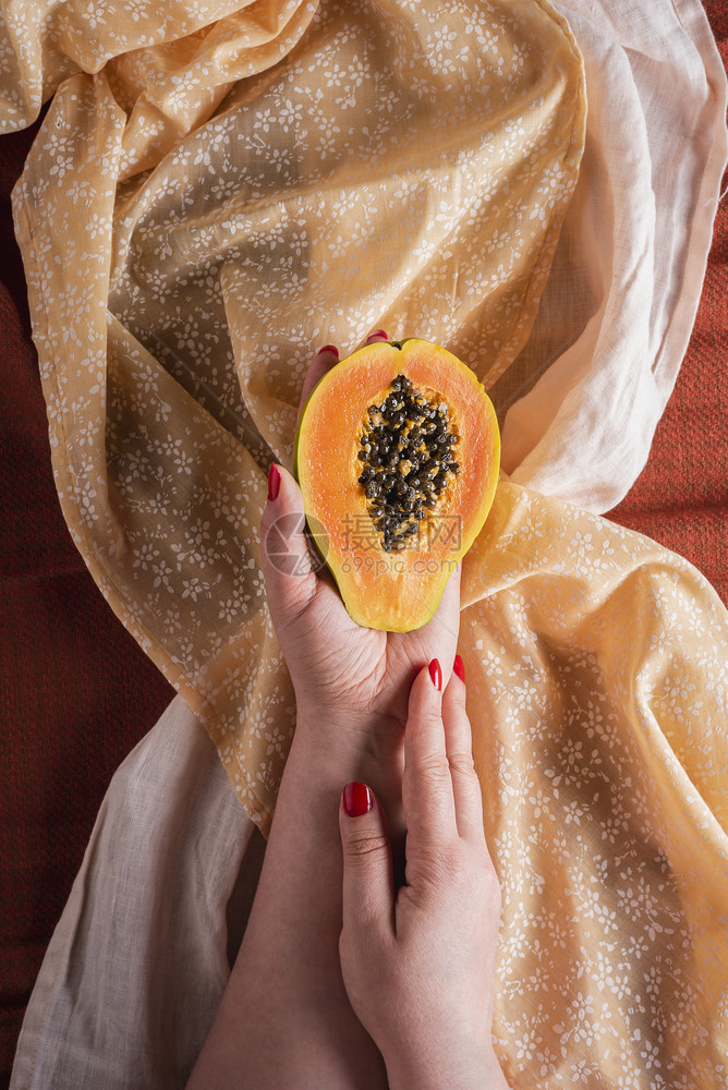 熟木瓜水果被切成两半在木瓜水果热带上方的黄色和橙桌布上由妇女用红钉子握在妇女手中图片