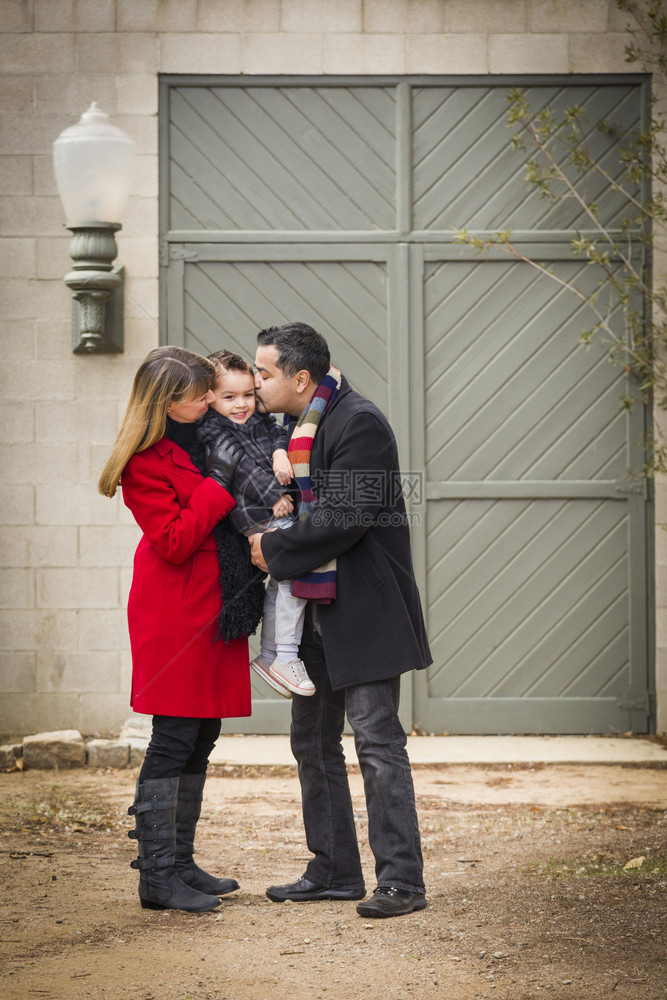 身着冬衣的年轻混血儿夫妇在新建筑前拥抱和亲吻儿子图片