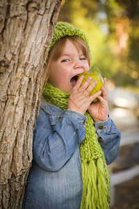 带着绿色围巾和帽子的可爱笑年轻女孩在外面吃着绿苹果图片