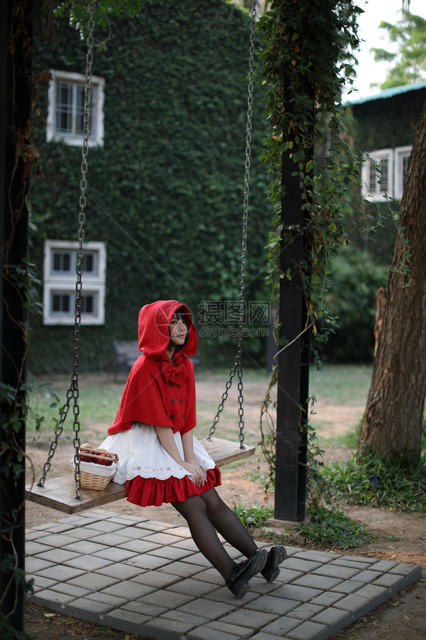 肖像身着小红色兜帽装饰的年轻女子在绿树公园背景的篮子上配有苹果和面包图片