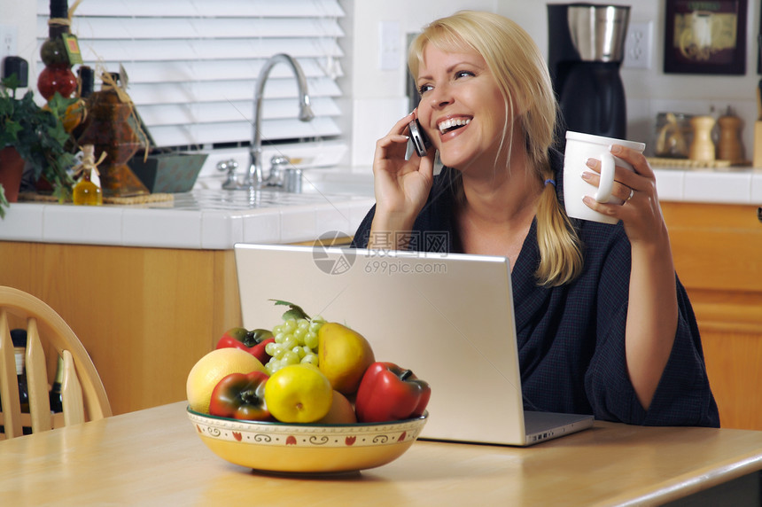 坐在膝上型电脑前的手机在厨房里微笑着妇女坐在厨房里的妇女用手机和膝上型电脑坐在厨房里的妇女图片