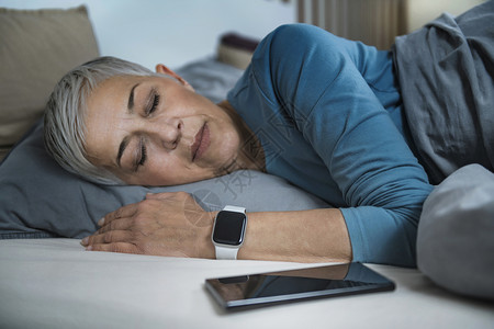 大咖睡眠应用高科技流经的老年妇女睡在床上使用智能电话和手表来改善她的睡眠习惯背景