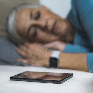 大咖睡眠应用高科技流经的老年妇女睡在床上使用智能电话和手表来改善她的睡眠习惯背景