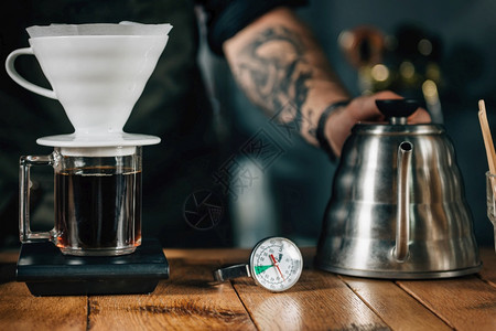 v字领贴上滴水咖啡机温度计和木制桌子上数字比例的瓶子制造滴水咖啡的工具和设备身穿黑制服的纹手臂咖啡师背景