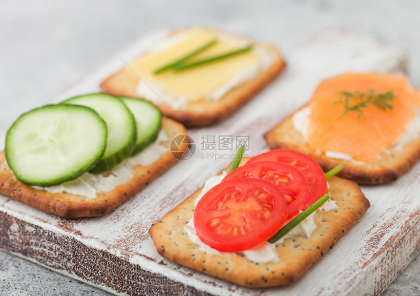 各种健康的饼干配有鲑鱼和奶酪番茄黄瓜放在浅色桌底的木切板上图片