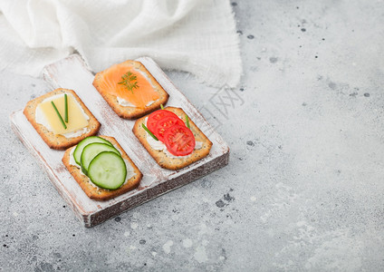 各种健康的饼干配有鲑鱼和奶酪番茄黄瓜放在浅色桌底的木切板上图片