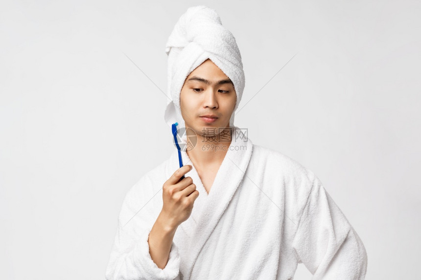 美丽spa和休闲概念穿着浴袍和巾的好奇深思熟虑的亚洲男子肖像看着对牙刷思考站在白色背景的思考中很感兴趣穿着浴袍和巾的好奇深思亚洲图片
