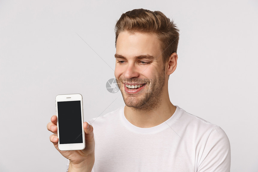 穿着白色t恤衫的可爱英俊金发长胡子男友拿着智能手机展示移动式微笑的喜悦吹嘘个人社交媒体页面申请评分喜悦的可爱英俊金发长胡子男友穿图片