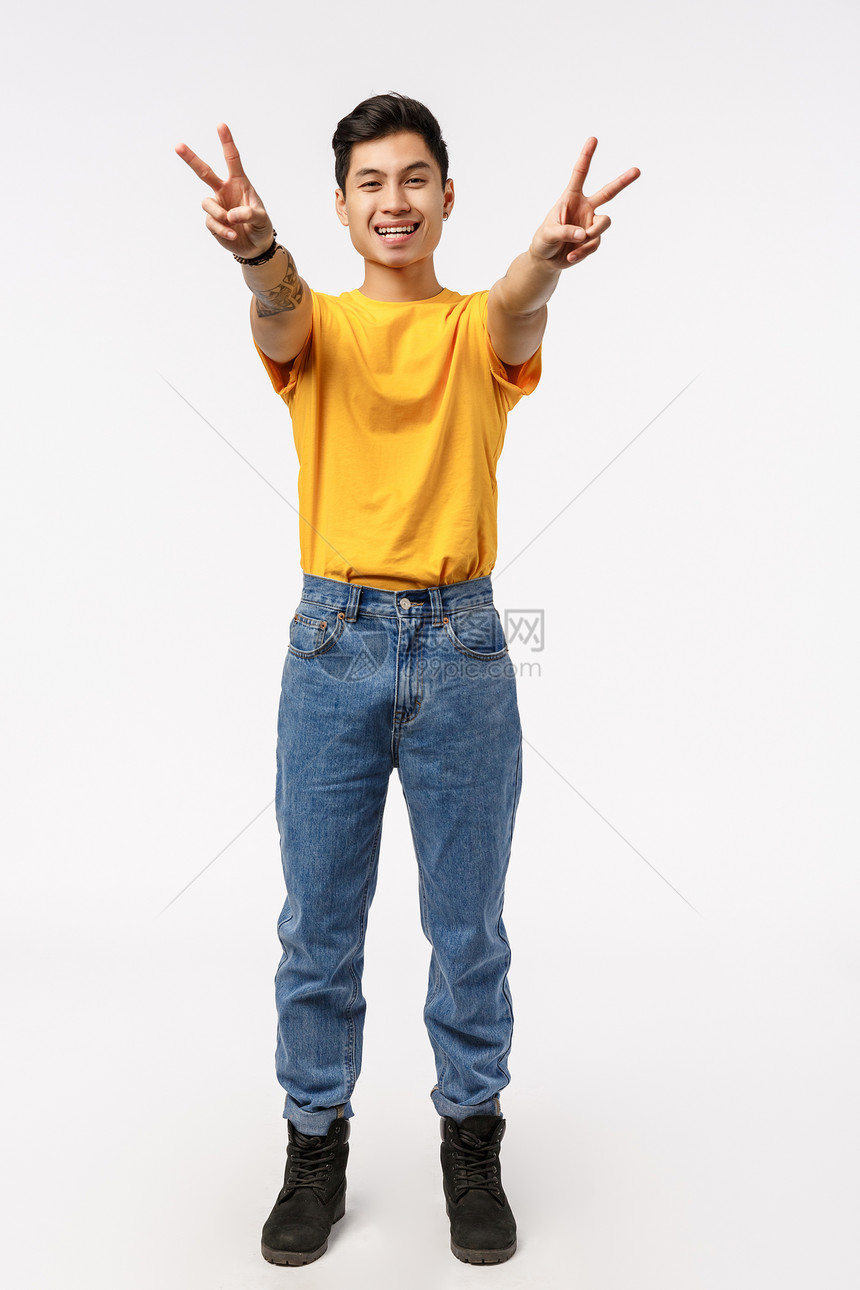 穿着牛仔裤黄色t恤衫展示和平或胜利迹象微笑表现实主义的快乐和无忧虑的情感向前手臂穿牛仔裤表现实主义的喜悦和无忧虑的情感纵向全拍的图片
