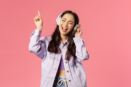 有吸引力的无照料亚洲女孩冷却和振动的肖像在耳机中听音乐在努力唱并达到歌的最高音调时举起手指粉红色背景有吸引力的无照料亚洲女孩冷却背景图片