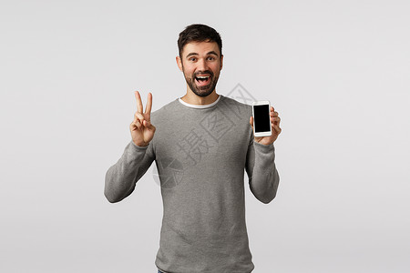 电话的标志表示和平标志展智能手机笑站立白色背景友善且自爱笑的男子在灰色毛衣编辑照片中制作和平标志笑站立白色背景背景