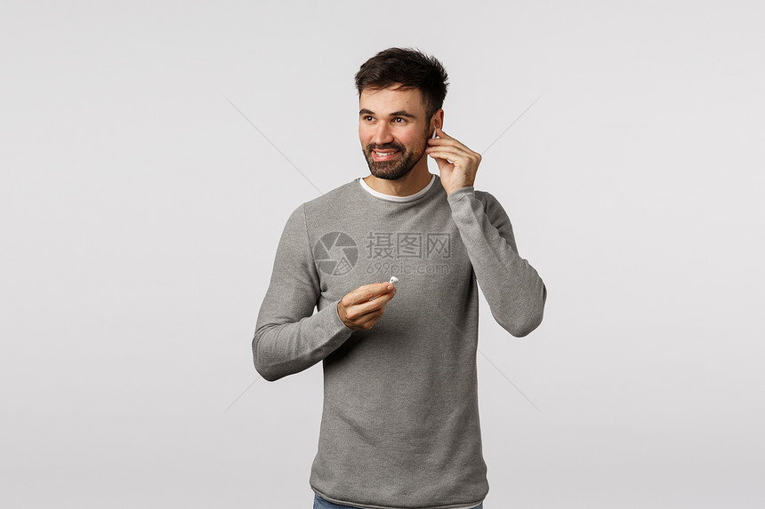 穿着灰色毛衣的有胡子成年男戴上耳机和微笑站在白色背景上欢乐男人准备出去戴上无线耳机来听音乐快的一天戴上耳机和微笑站在白色背景上欢图片