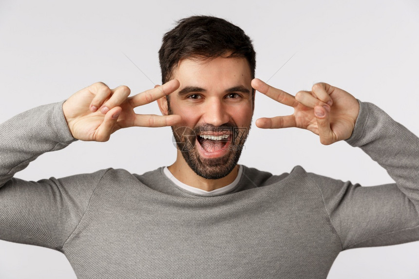 有胡子的快乐男子穿着灰色毛衣展示迪斯科的姿态和平标志在眼睛上微笑欢呼起来玩得开心在节假日祈祷做个好交易图片