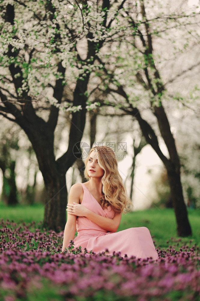 花朵中美丽的金发女在开花的春季公园中有化妆品和发型的粉红色裙子女孩妇和一天花朵中有美丽的金发女图片