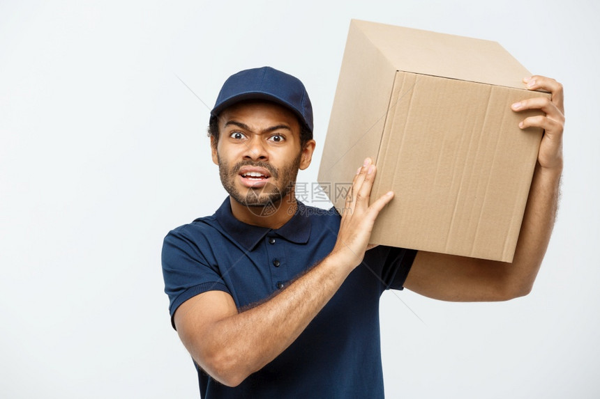 交付概念严肃的非洲送货员肖像显示愚蠢的攻击表达方式持有一个盒子包孤立在灰色工作室背景上复制空间图片