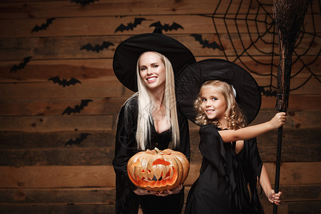巫家坝halowen概念快乐的母亲和女儿穿着巫服庆祝halowen装扮有弯曲南瓜的圣殿在木制工作室背景上的蝙蝠和蜘蛛网之上打扮成弯曲南背景