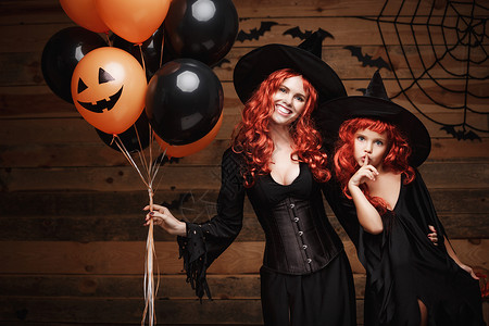 欢乐的母亲和她女儿穿着巫服装庆祝圣殿扮成橙色和黑的气球在木制工作室背景上披橙色和黑的气球蜘蛛网欢乐的母亲和她女儿穿着巫装背景图片