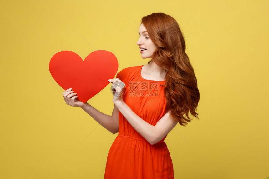 年轻快乐的红发女郎穿着橙色的漂亮裙子,穿着大红心纸。 生活方式和节日概念,年轻快乐的红发女郎穿着大心纸。图片