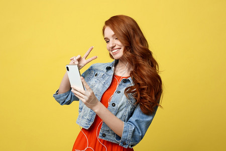 生活方式概念用手机为穿散装的姜发妇女肖像用手机为互动视频打电话显示两只手指在明黄色背景上隔离生活方式概念用手机为穿散装的姜发妇女图片