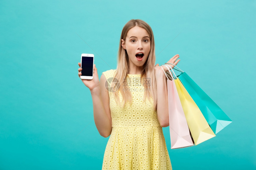 带有购物袋的吸引力年轻女肖像显示手机和屏幕直接到相机上以蓝色背景隔离带购物袋的有吸引力年轻女肖像显示手机和屏幕直接到相机上隔离图片