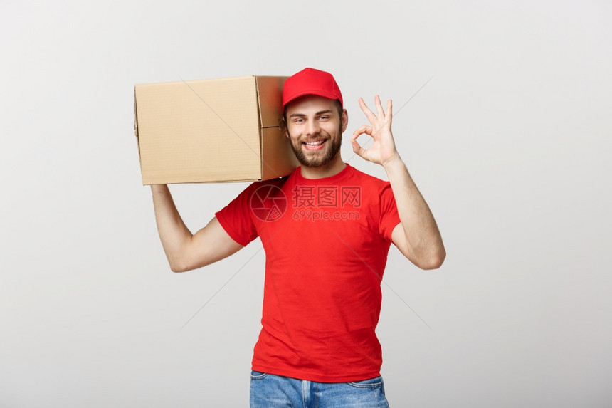 快乐的年轻送货员站在红帽子上白背景隔开的邮箱表现不错快乐的年轻送货员站在红帽子上包裹邮箱在白色背景上隔开表现不错的手势图片