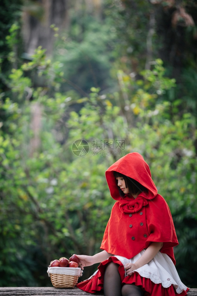 肖像身着小红色兜帽装饰的年轻女子在绿树公园背景的篮子上配有苹果和面包图片