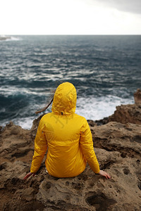 黄色的雨衣身着黄色雨衣坐在悬崖上看着海浪的大同时也享受着美丽的海景在雨天中下云雾的春季天气中岩石海滩上背景