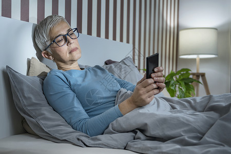 睡不着的成年妇女睡不着在床上网用智能手机通过社交网络给朋友发短信图片