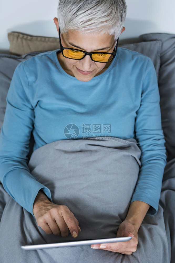 身戴蓝灯眼镜黄睡前躺在床上看着平板屏幕的成年妇女图片