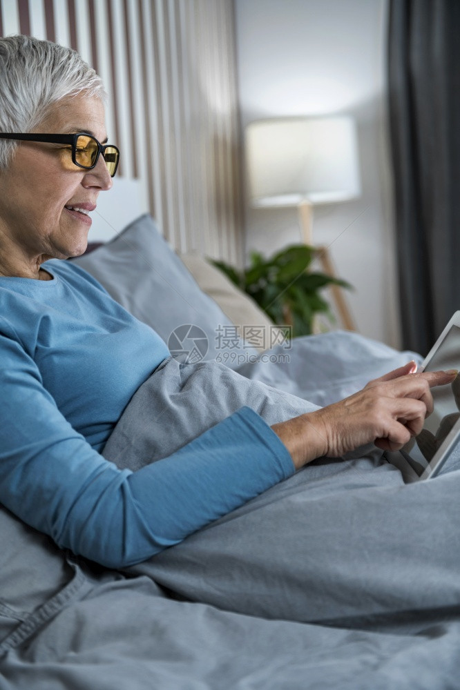 身戴蓝灯眼镜黄睡前躺在床上看着平板屏幕的成年妇女图片