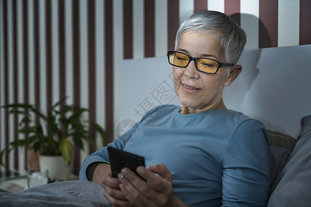 睡前用智能手机把眼镜放在床上晚用智能手机把眼镜用蓝色灯挡住的老年妇女图片