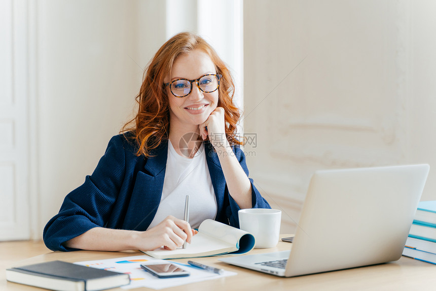 成功的眼衣经济学家检查膝上型计算机的信息在金融网站上阅读信息用笔记本写作喝热咖啡坐在工作场所图片