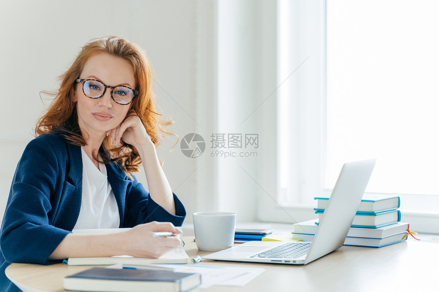 成功的红头女企业主自信的横向拍摄坐在内阁里笔记本电脑上工作在笔记本里写主要论文喝咖啡休息在宽敞的办公室里化妆图片