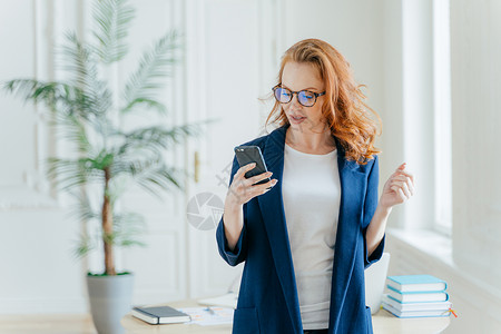 成功女商人在手机上使用应程序阅读老板或同事的回信电子邮件身着正式服装在办公室内部摆姿势发现金融新闻背景