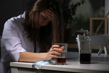 疲劳的女医生坐在桌子上辛勤工作之后在家中用威士忌或白兰地杯子拿起威士忌或白兰地抑郁的女医生喝着有冠状腺问题的烈酒精疲劳的女医生坐背景图片
