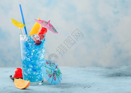 高球玻璃中的蓝色环礁湖夏季鸡尾酒配有甜樱桃和橙色切片蓝桌底有雨伞背景图片