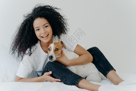 快乐的阿夫罗女人与狗同床共枕心情有趣与白色背景一起在卧室里摆姿势女孩在家和杰克罗瑟尔特里一起放松背景图片