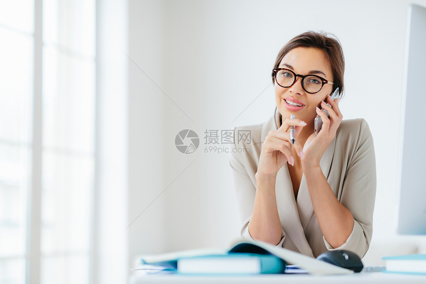 女雇员的照片有快乐表情通过智能手机交谈握笔坐在桌面上有纸张和笔记本计算机反办公室内部的姿势佩戴眼镜优雅的衣服图片