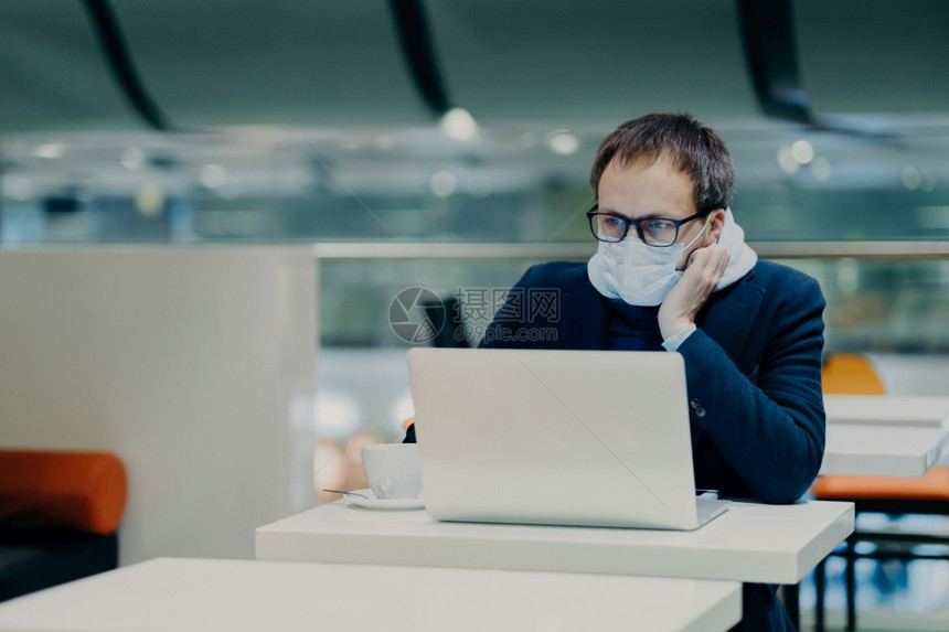 重人照片集中在笔记本电脑屏幕上在冠状爆发时从远处工作戴防护面具不传播疾病喝咖啡坐在白桌旁图片