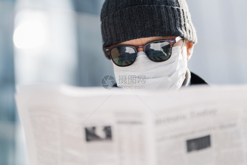 近距离拍摄的成年男子戴太阳镜帽和无菌的医学面具阅读报纸发现世界形势的消息传出冠状在模糊的背景之下在户外传播图片