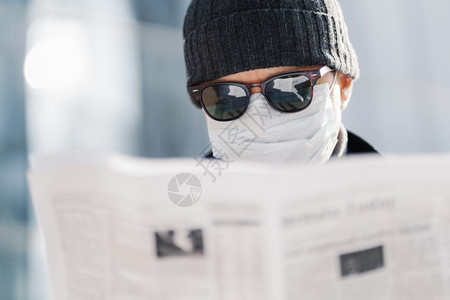 近距离拍摄的成年男子戴太阳镜帽和无菌的医学面具阅读报纸发现世界形势的消息传出冠状在模糊的背景之下在户外传播背景图片