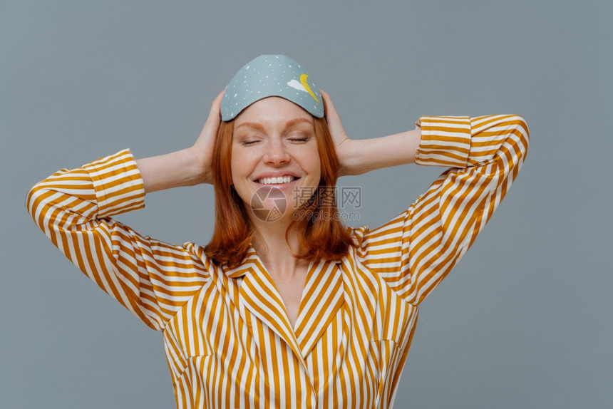 快乐的有姜长发女人穿着舒适脱条睡衣和面具使用有的辅助剂睡觉笑得面带微展示白牙在灰色工作室墙壁上隔离图片