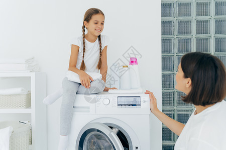 自助小料可爱的小女孩有猪尾辫在洗衣机顶上摆着姿势拿白色软毛巾高兴地看着母亲在洗完衣服后谈计划背景