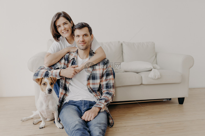 喜悦的女子相片拥抱她的丈夫在沙发上摆姿势小狗坐在地上靠近房主图片